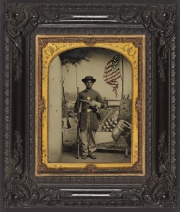 Unidentified African American soldier in Union uniform with a rifle and revolver in front of painted backdrop showing weapons and American flag at Benton Barracks, Saint Louis, Missouri (by Enoch Long, between 1863 and 1865; LOC: LC-DIG-ppmsca-36456)