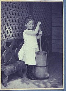 Little girl wearing white chemise, beaded necklace, and high-button shoes on porch with butter churn (ca1900; LOC: LC-USZC2-6076)