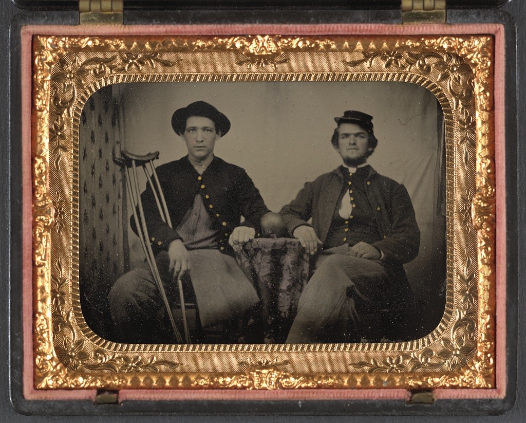 Two unidentified soldiers in Union private's uniforms sitting next to table with cannon ball on top; one soldier has an amputated leg and holds crutches (between 1861 and 1865; LOC: http://www.loc.gov/item/2012648229/)