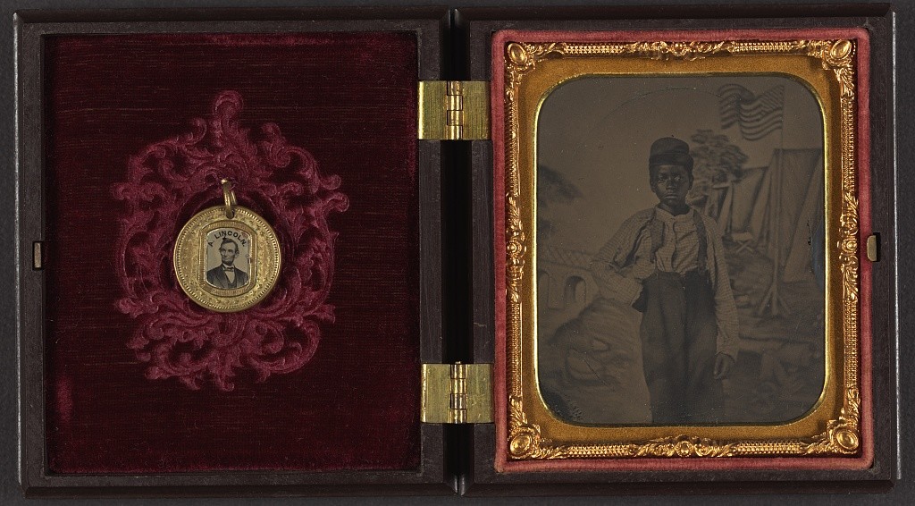 Unidentified African American boy standing in front of painted backdrop showing American flag and tents ; campaign button with portraits of Lincoln on one side and Johnson on the opposite side are attached to inside cover of case (between 1861 and 1865; LOC: http://www.loc.gov/item/2010648377/)
